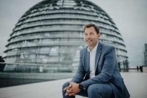 SPD Mitglied Lars Klingbeil sitzt vor der Kuppel des Bundestages. Er trägt einen grauen Anzug und ein weißes Hemd.