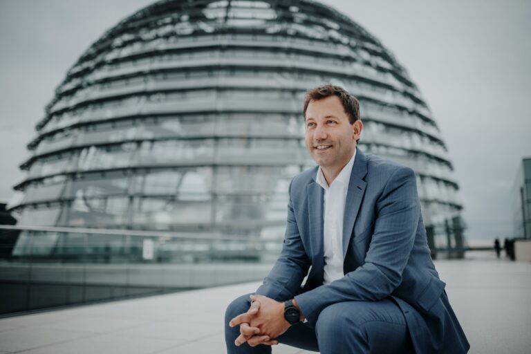 SPD Mitglied Lars Klingbeil sitzt vor der Kuppel des Bundestages. Er trägt einen grauen Anzug und ein weißes Hemd.