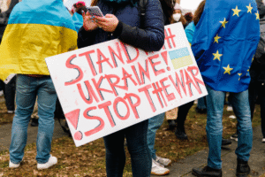 Menschen bei einer Demonstration. Ein Mann hält ein Handy in der Hand. Unter seinem Arm hat er ein Schild geklemmt, auf dem in roten Buchstaben zu lesen ist: Stand with Ukraine. Stop the War.