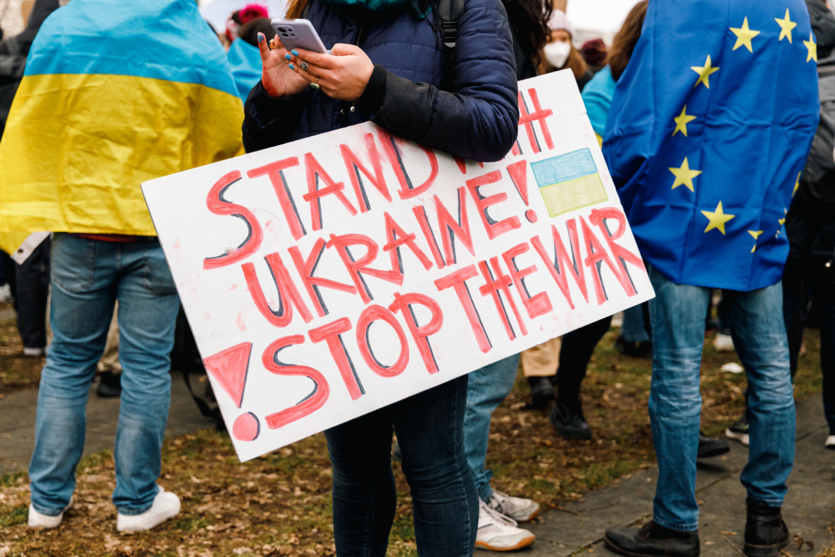 Menschen bei einer Demonstration. Ein Mann hält ein Handy in der Hand. Unter seinem Arm hat er ein Schild geklemmt, auf dem in roten Buchstaben zu lesen ist: Stand with Ukraine. Stop the War.